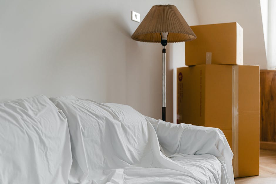 Inside a home during a furniture transport process, four cardboard boxes are stacked near a wooden cabinet against a beige wall. The boxes are sealed with packing tape and vary in size. To the left, a